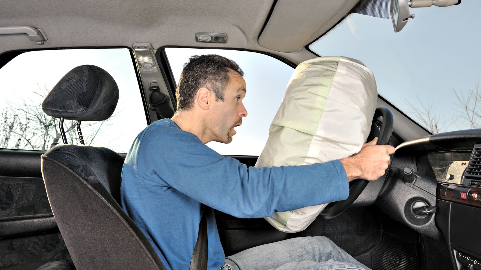 a man sitting in a car holding a pillow.|a steering wheel with a cloth on it.|a car with a bunch of items in the passenger seat.