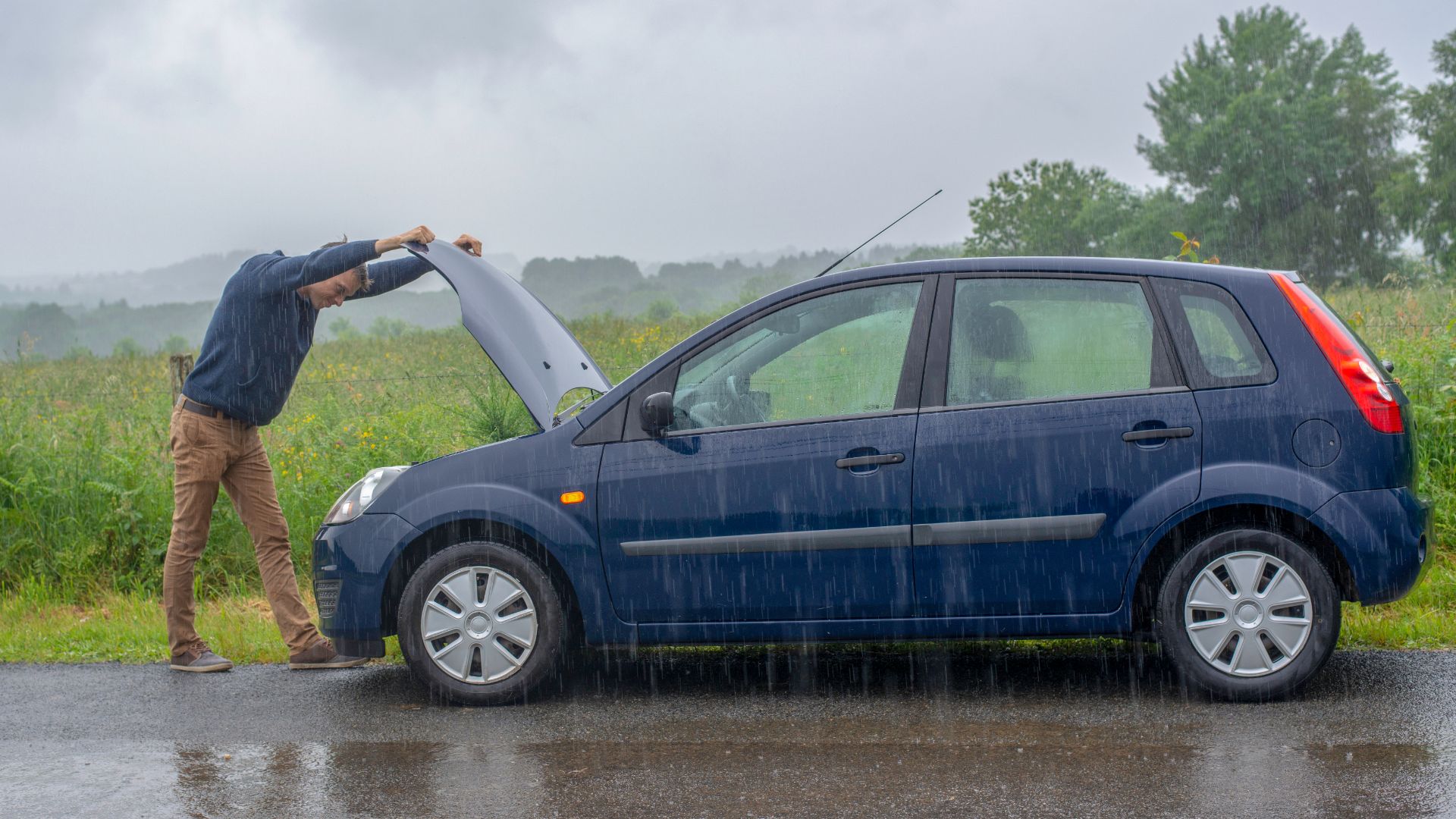 a man standing next to a blue car in the rain.|a pair of pliers are hooked up to a car battery.