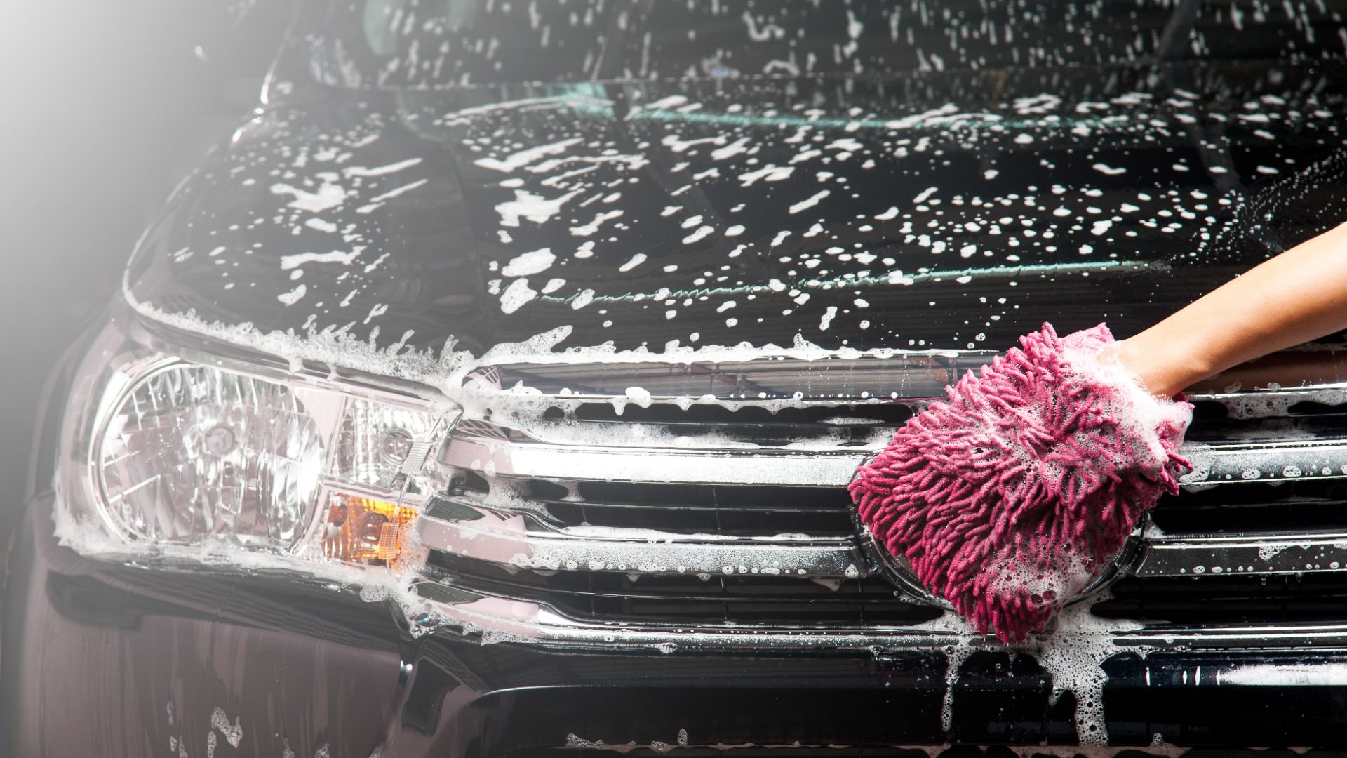 a person with a pink mitt on top of a black car.|a person washing a car with a rag.|a close up of a car with a lot of water on it.