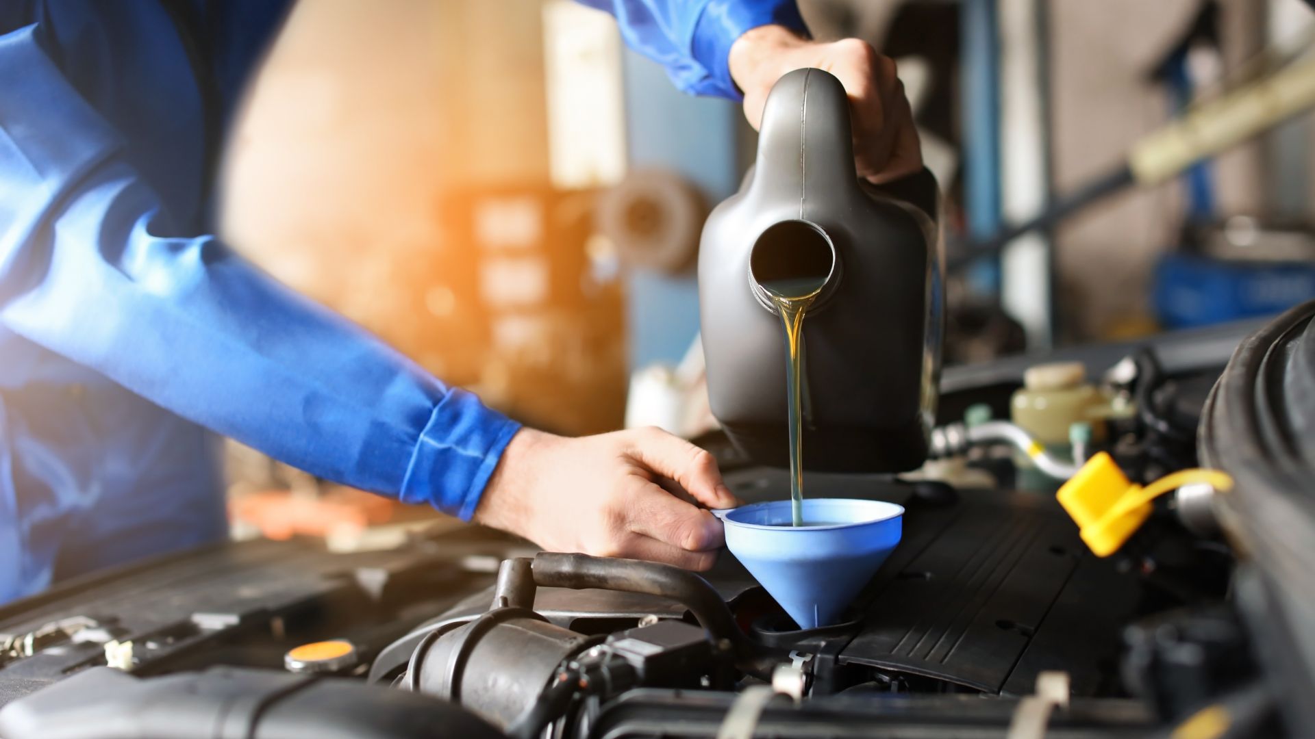 a man pouring oil into a blue bowl.|two men working on a car in a garage.