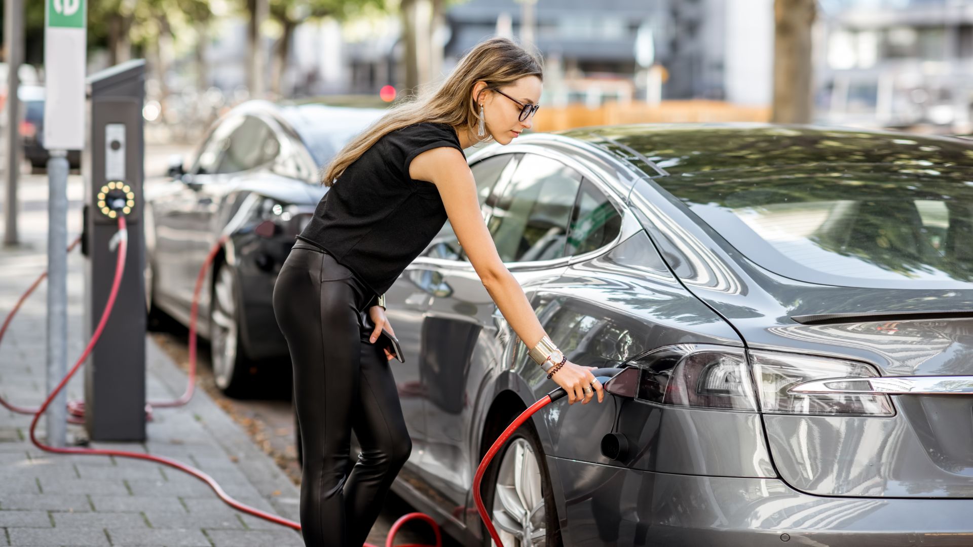 a woman is charging her car on the street.|a couple of people that are looking at a machine.