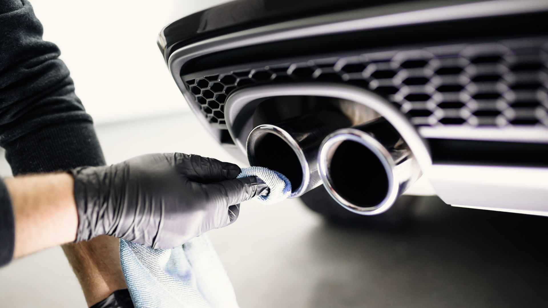 a person wearing gloves and gloves is cleaning the front end of a car.|a mechanic working on a car in a garage.|a close up of a car's exhaust system.