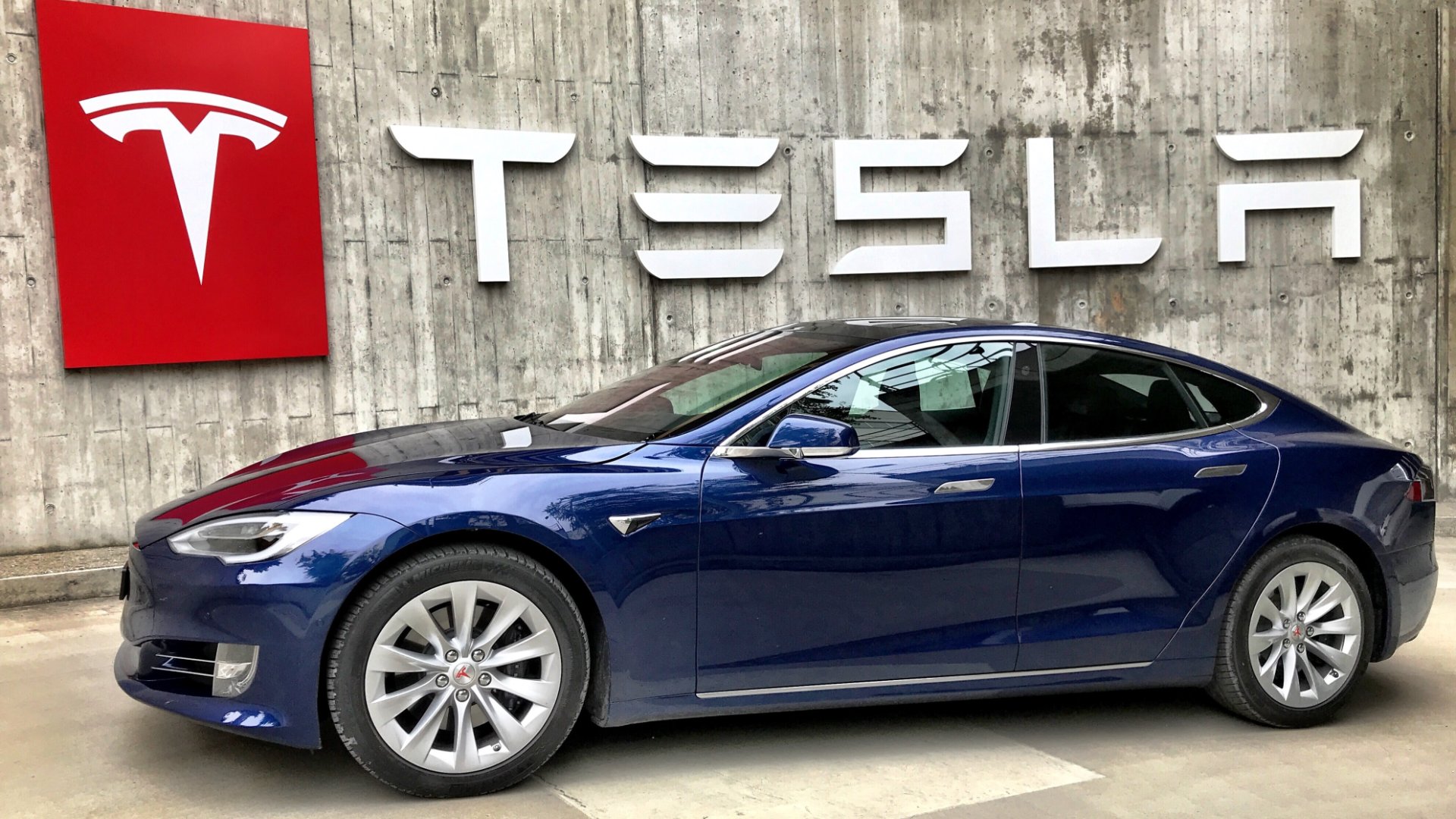 a blue tesla car parked in front of a tesla sign.|a blue tesla electric car at a gas station.