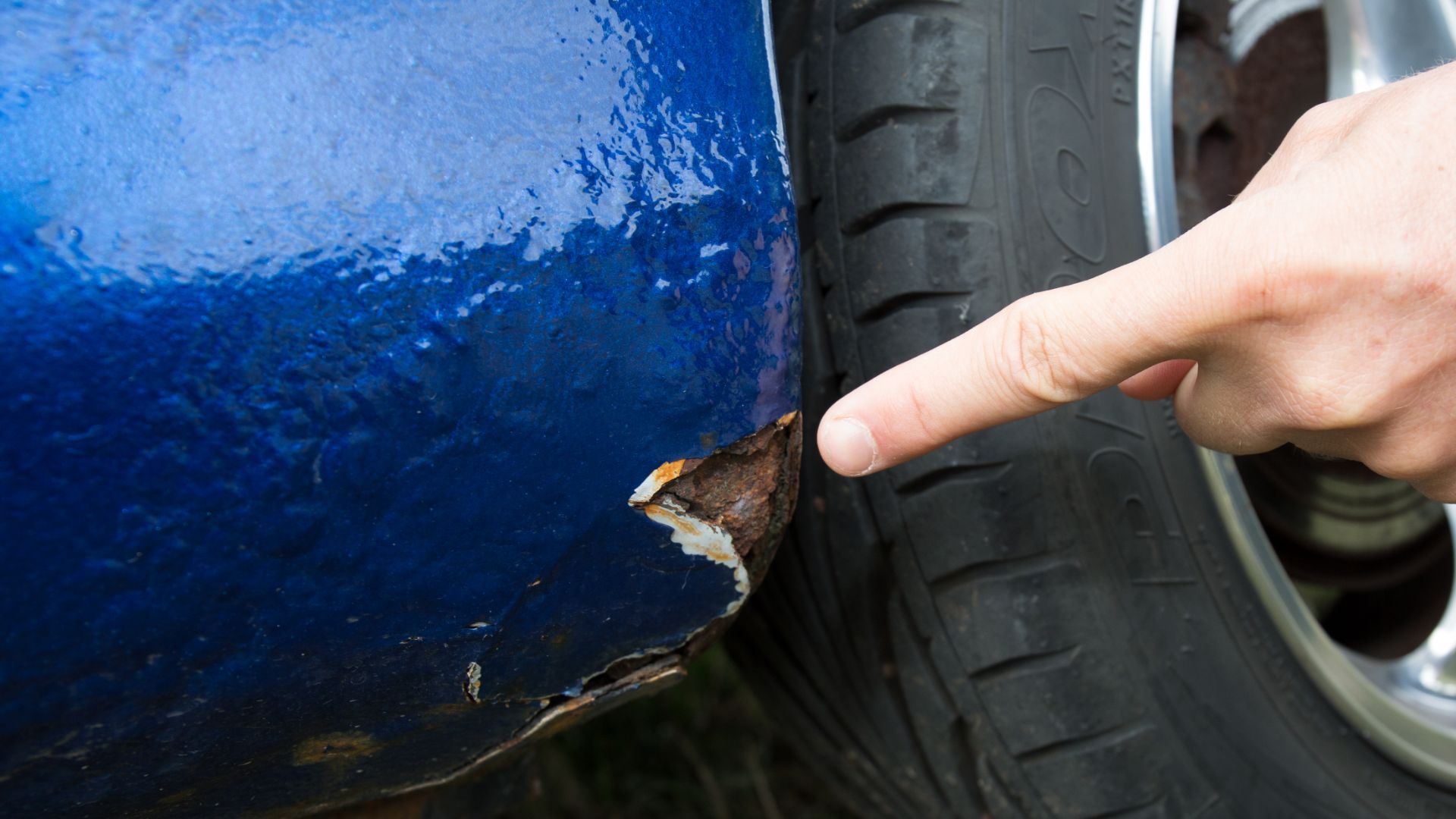 a person points at a piece of rust on the side of a blue car.|a close up of the front of a car with rust on it.|a close up of a tire with rust on it.|a close up of the side of a red car.|a person cleaning a car with a blue cloth.|a close up of the side of a car with a rusted tire.